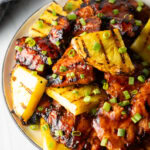 Top down view of sticky chicken pieces topped with chopped scallions and grilled slices of pineapple.