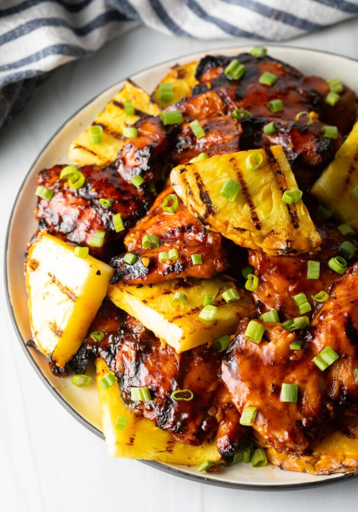 Top down view of sticky chicken pieces topped with chopped scallions and grilled slices of pineapple.