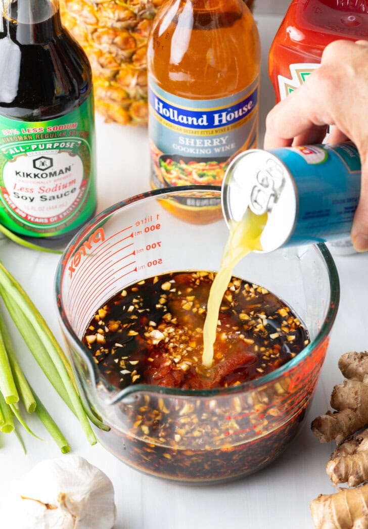 Hand adding pineapple juice from a can into a huli huli chicken marinade in a large glass measuring cup.