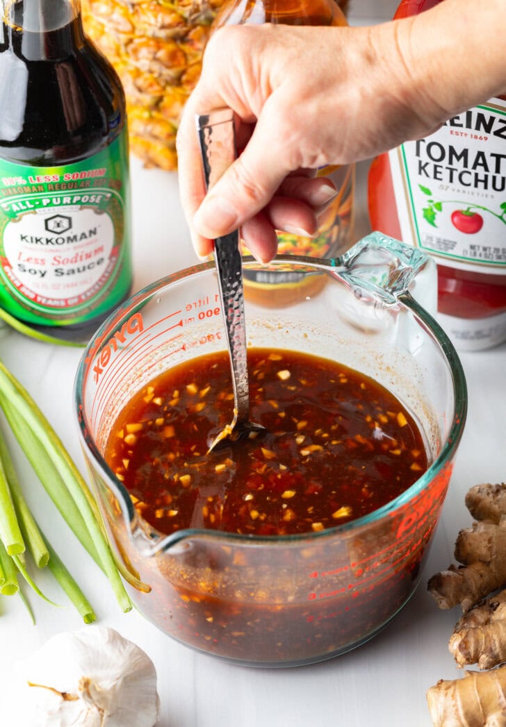 Hand using metal spoon to stir the Hawaiian chicken marinade in a large glass measuring cup.