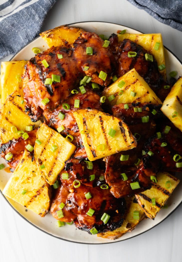 Top down view of sticky chicken pieces topped with chopped scallions and grilled slices of pineapple.