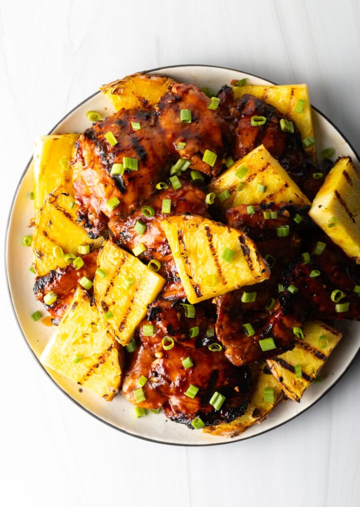 Top down view of sticky chicken pieces topped with chopped scallions and grilled slices of pineapple.