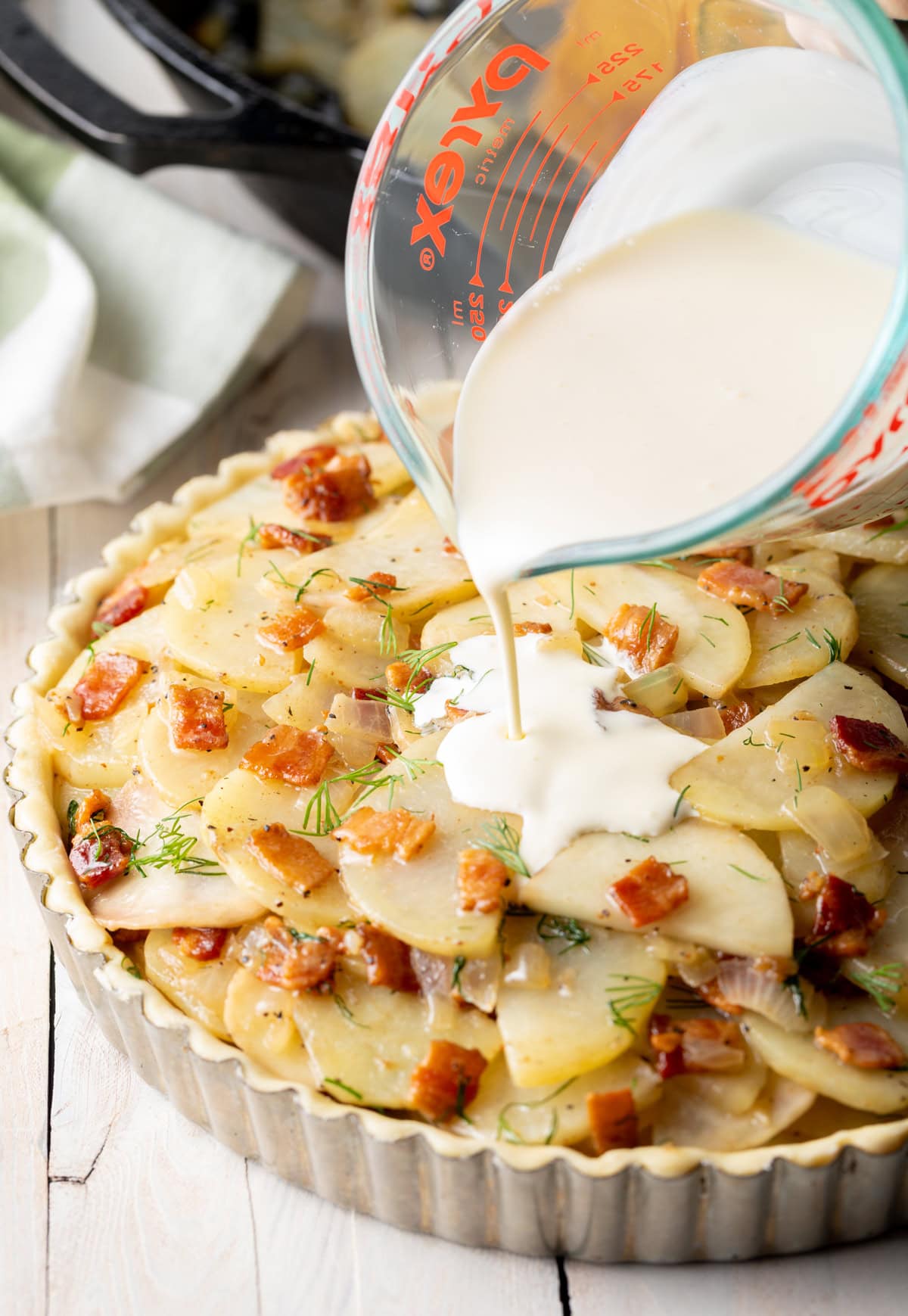 Heavy cream being poured over the top of potatoes and bacon in a tart tin. 