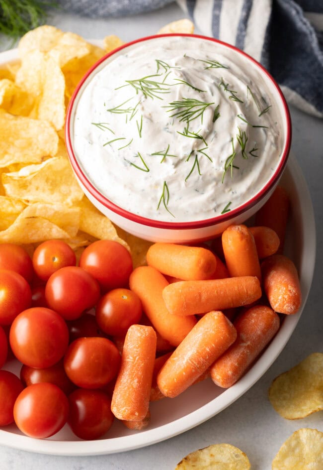 Top view bowl of dip with fresh chopped dill on top. Bowl is nestled on a plate with baby carrots, cherry tomatoes, and potato chips.