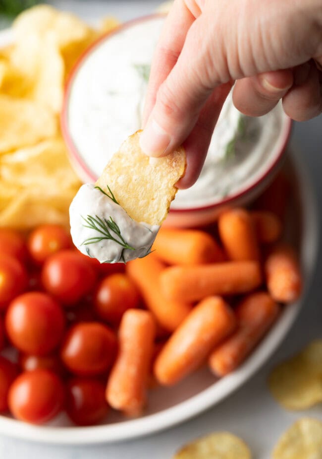 Hand with potato chip and dill dip, shown to camera hovering above plate with cherry tomatoes, baby carrots, and potato chips.