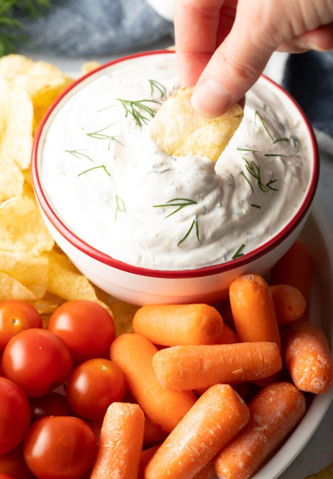 Hand dipping potato chip into bowl of creamy dill dip.