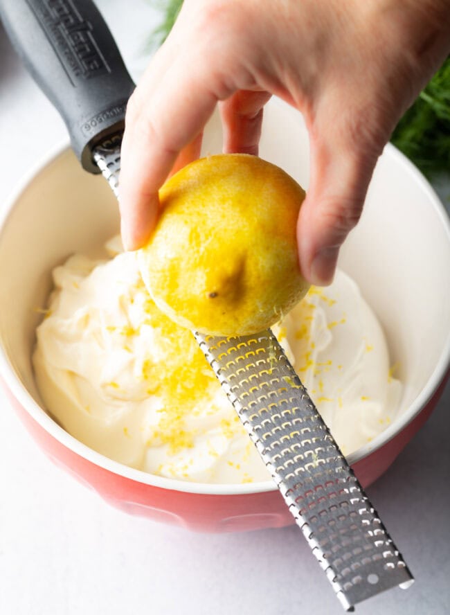 Hand zesting lemon over bowl of sour cream.