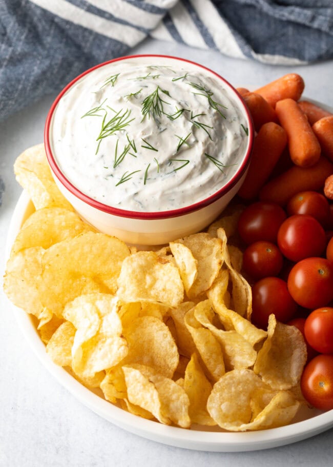 Creamy sour cream dip in a bowl, surrounded by potato chips, cherry tomatoes, and baby carrots.