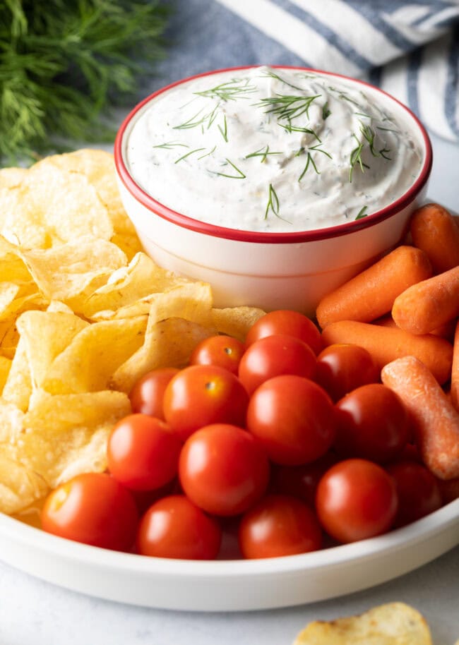 Dip in a bowl, surrounded by potato chips, cherry tomatoes, and baby carrots.