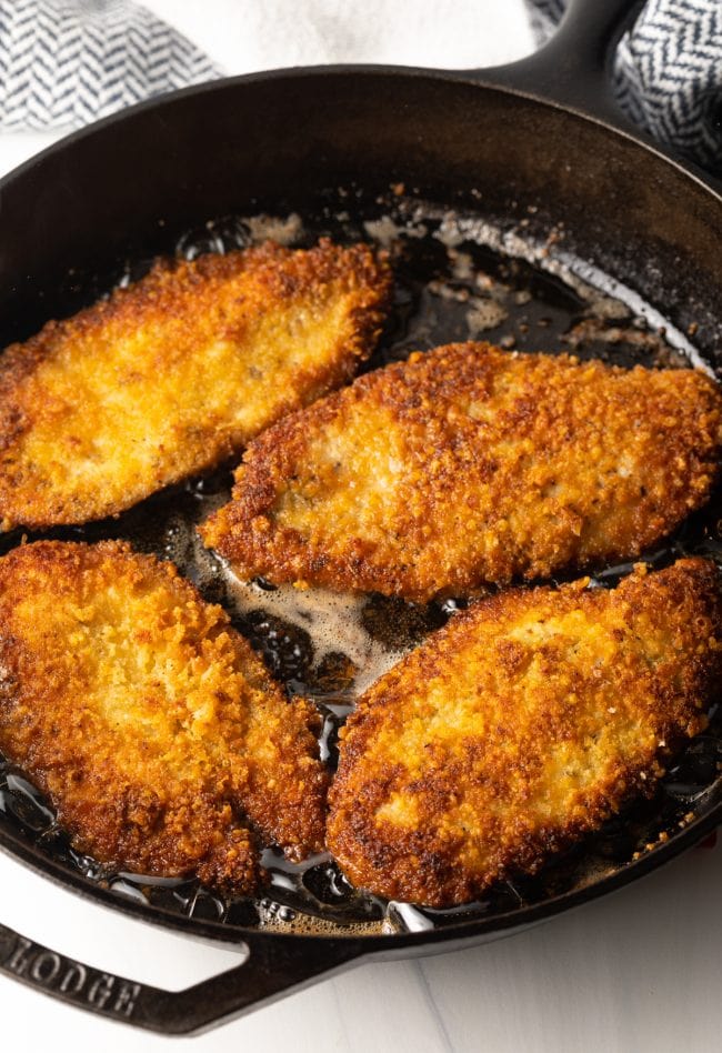 large cast iron pan frying crispy chicken cutlets with Italian marinade and seasoning