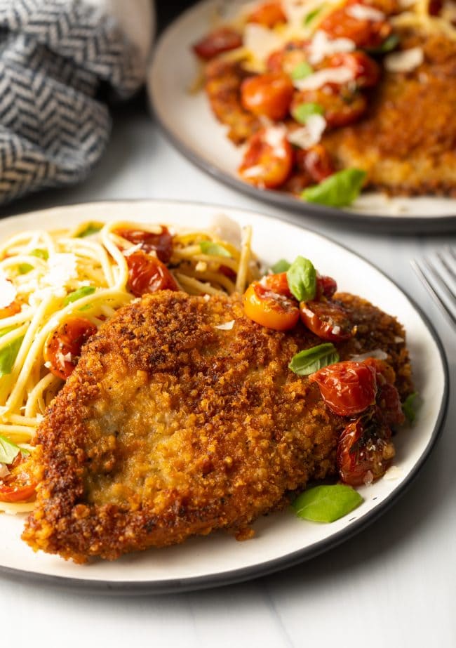 side view breaded fried Italian chicken cutlets on a plate with pasta and tomatoes and micro basil