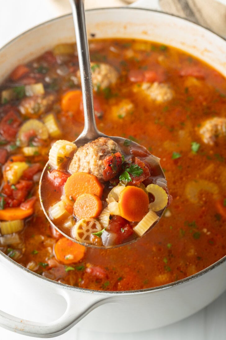 Metal ladle holding serving of soup to camera. Large white pot of soup is in the background.