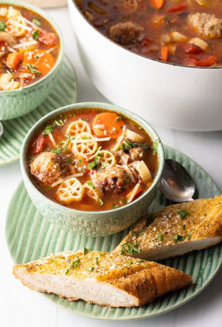 Green bowl of Italian meatball soup loaded with wheel shaped pasta, sliced carrots, in a tomato broth. Two pieces of toasted baguette are on the green plate that's beneath the soup.