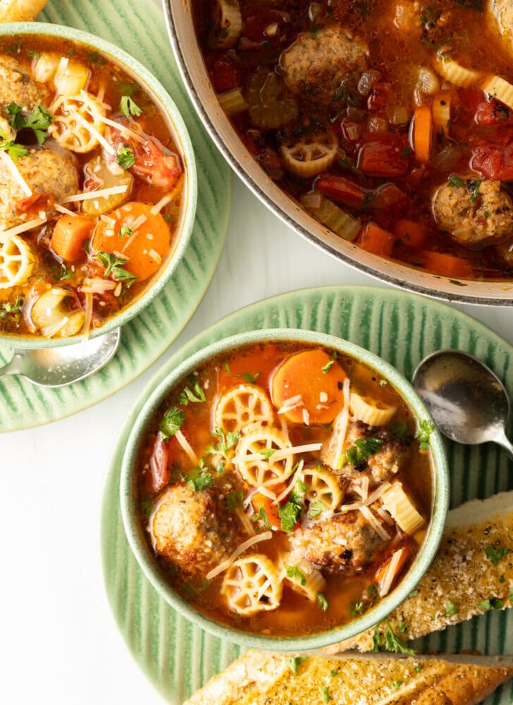 Top down two green bowls of Italian meatball soup loaded with wheel shaped pasta, sliced carrots, in a tomato broth.