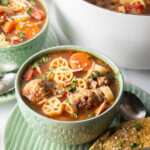 Green bowl of Italian meatball soup loaded with wheel shaped pasta, sliced carrots, in a tomato broth.