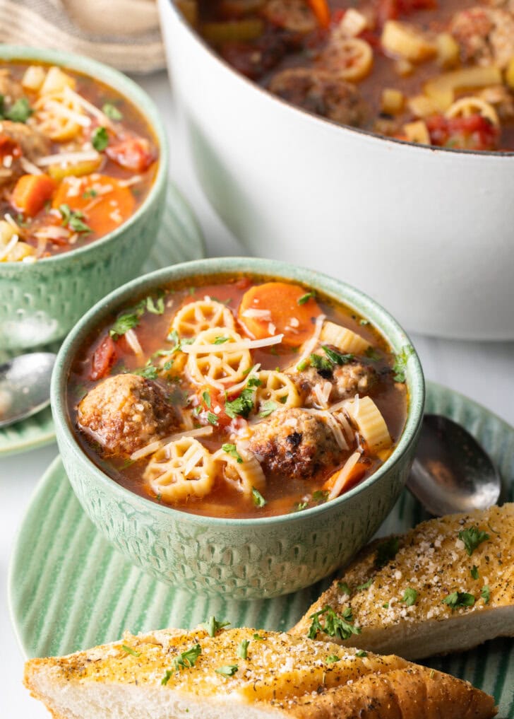 Green bowl of Italian meatball soup loaded with wheel shaped pasta, sliced carrots, in a tomato broth.