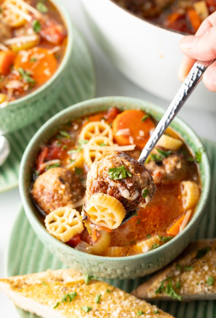 Green bowl of Italian meatball soup loaded with wheel shaped pasta, sliced carrots, in a tomato broth. A metal spoon is holding a bite up to camera.