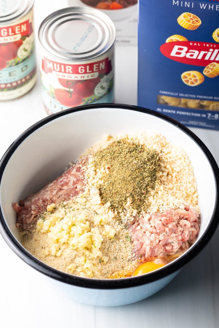 Meatball ingredients in a large white mixing bowl: ground sausage, breadcrumbs, diced onion, and spices.