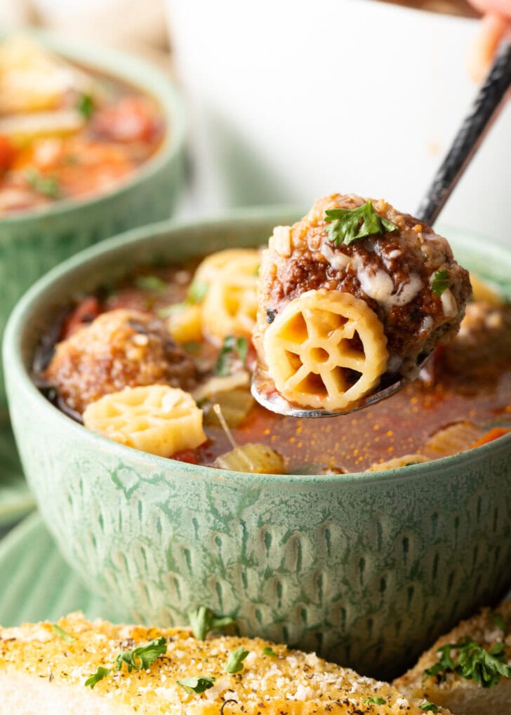 Green bowl of Italian meatball soup loaded with wheel shaped pasta, sliced carrots, in a tomato broth. A metal spoon is holding a bite up to camera.