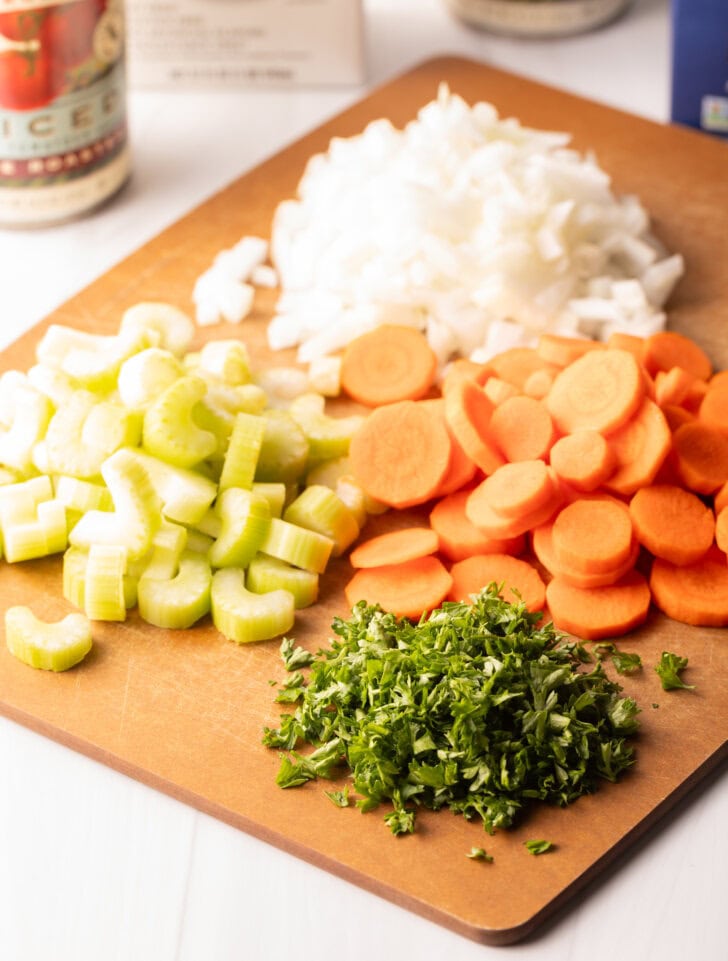 Wooden cutting board with individual piles of slices carrots, celery, diced onion, and parsley.