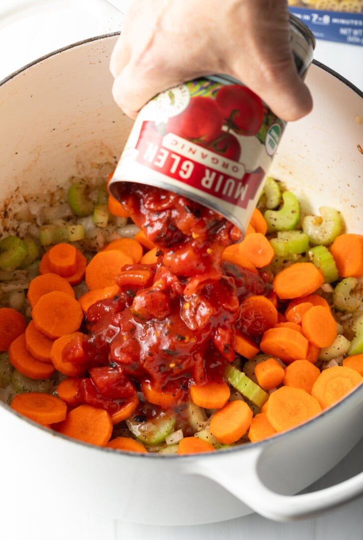 Hand adding canned tomatoes to pot with fresh slices of carrots and celery.