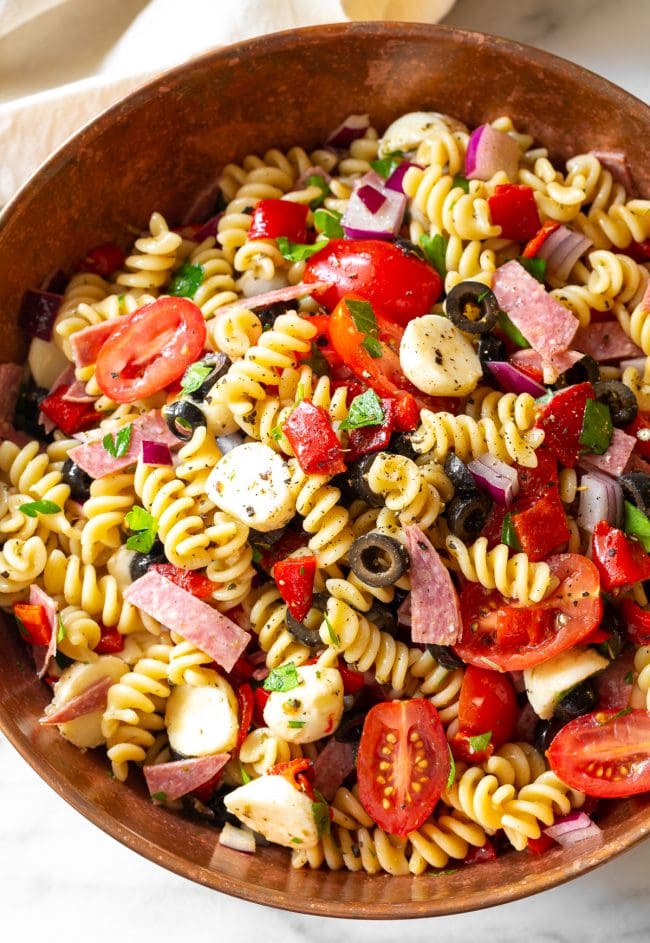 Noodles, veggies, salami, cheese and olives with dressing in a large bowl. 