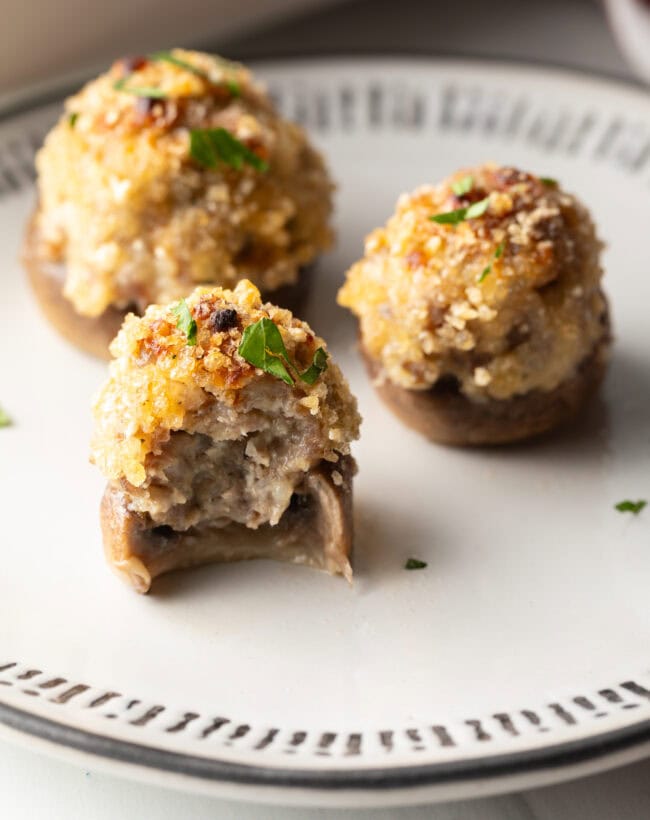 Three mushrooms stuffed with Italian sausage and cheese on a white plate. Front mushroom has been bitten in half to show inside.