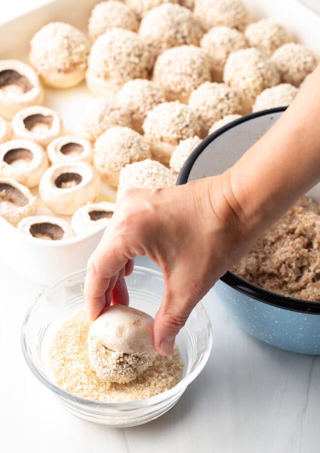 Hand holding a stuffed mushroom cap above a bowl of breadcrumbs.