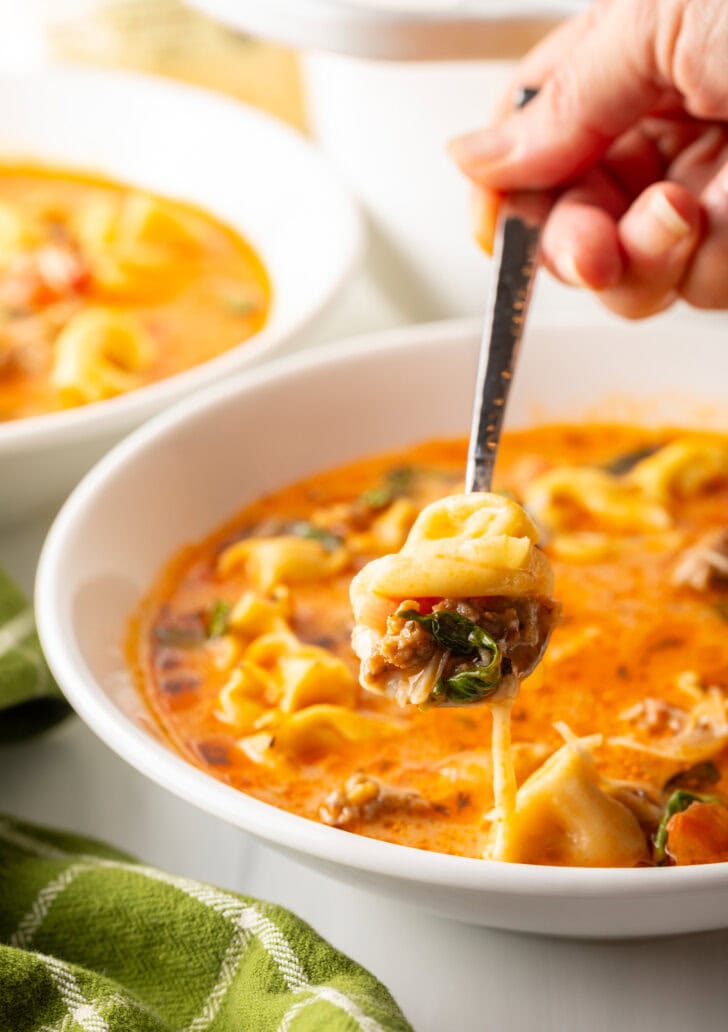 Hand with metal spoon, scooping a bite of tortellini and basil from a bowl of soup and showing to camera.