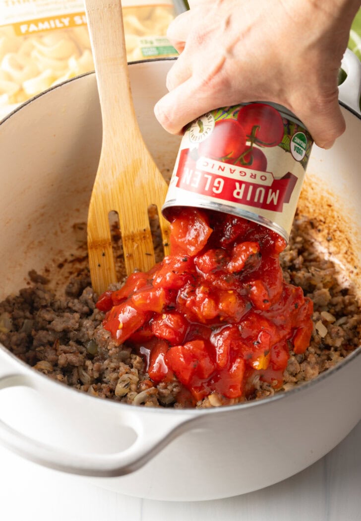 Hand pouring diced tomatoes from a can into the soup pot with ground sausage and wooden spatula.