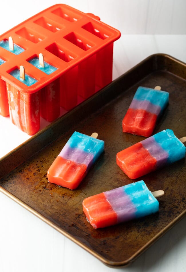 4 rainbow popsicles on a rimmed baking sheet, ice pops in mold in background.