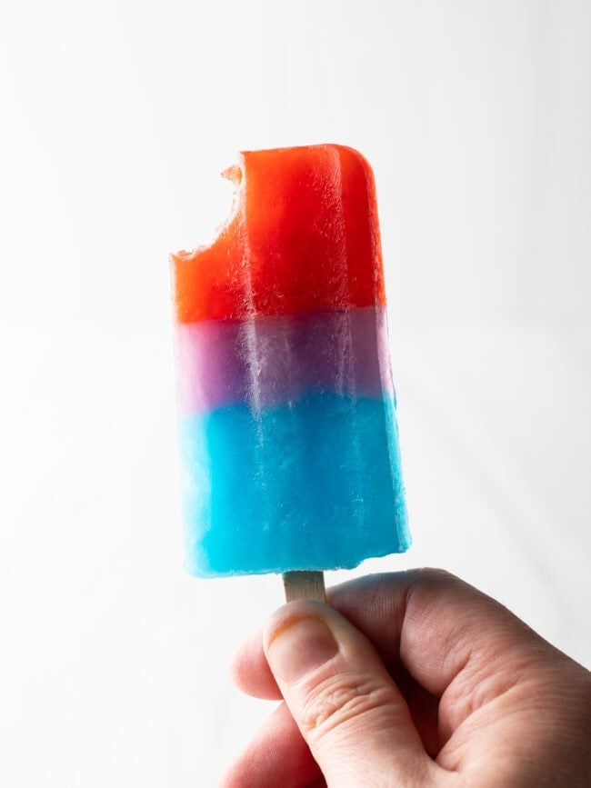 Hand holding up red, purple, blue popsicle with bite taken out of top red section.