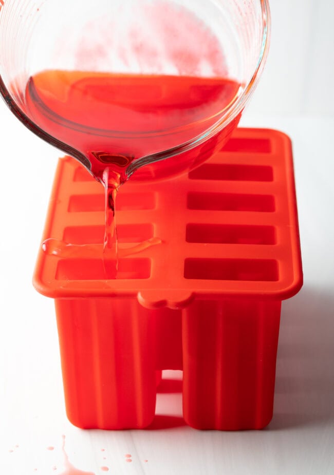 Glass measuring cup pouring red liquid into red popsicle mold.