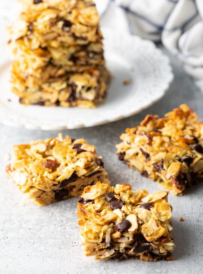 Several squares of cereal treats with coconut and nuts on a gray background.