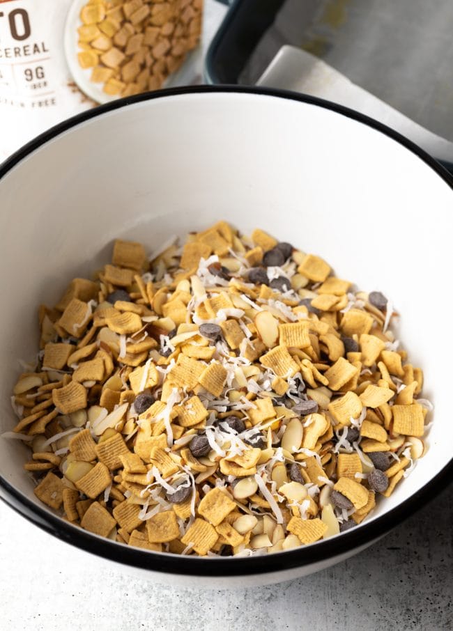 Cereal, chocolate chips, shredded coconut, and nuts in a large white mixing bowl.