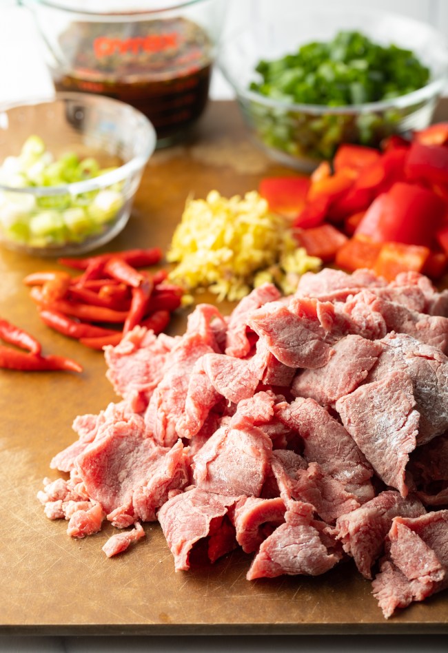 Recipe ingredients chopped on a cutting board in individual piles: steak, onion, red bell peppers, garlic.