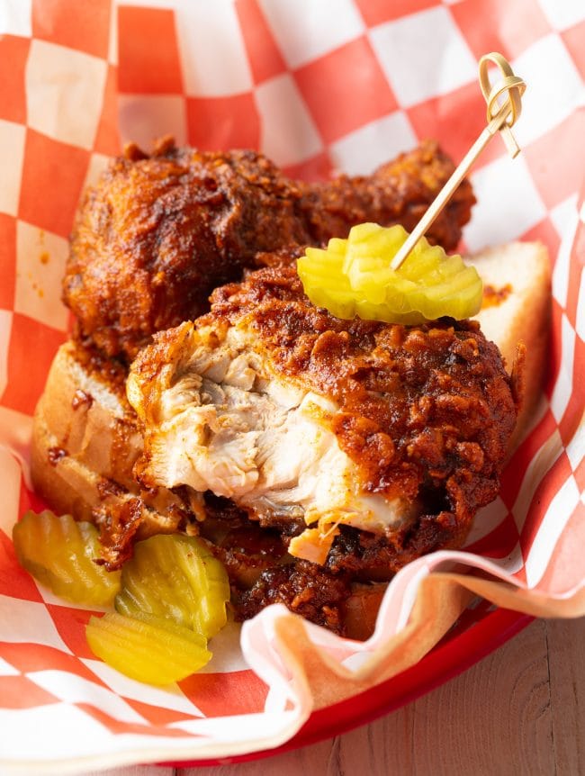 Spicy fried chicken cut open to expose juicy breast, in a serving basket with pickles. 