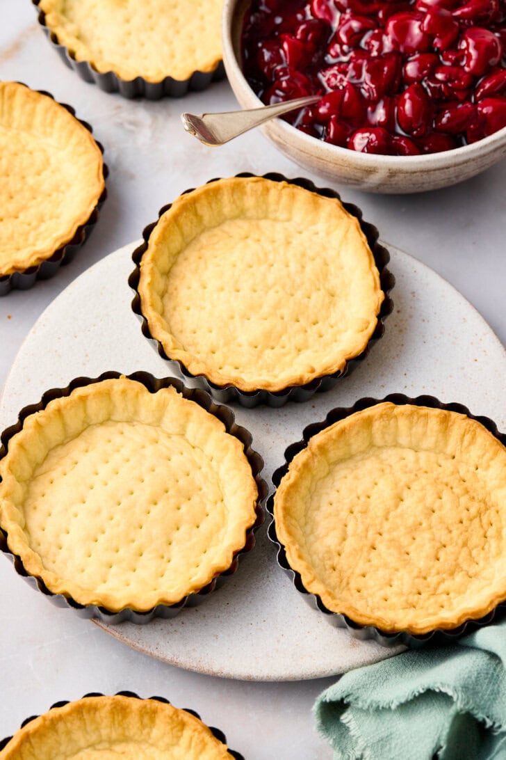 Baked cherry pie crusts in the tart pans next to a bowl of cherry pie filling. 