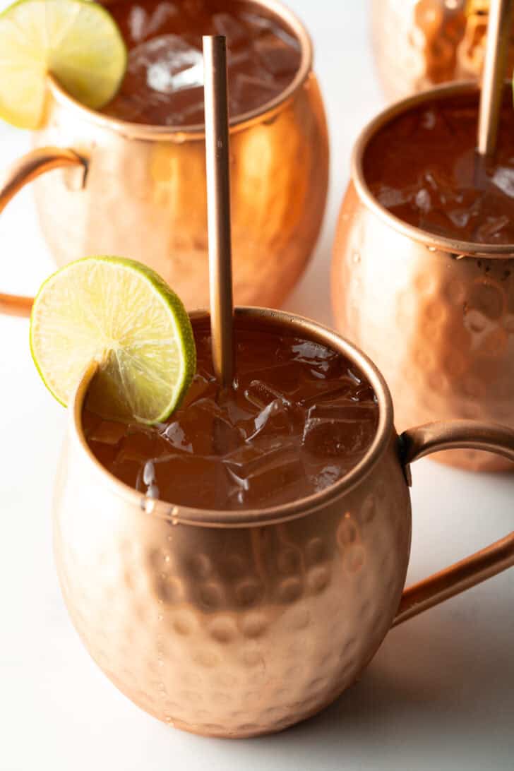 Close view of a moscow mule in a copper handled mug, with a copper metal straw and slice of lime to garnish the rim.