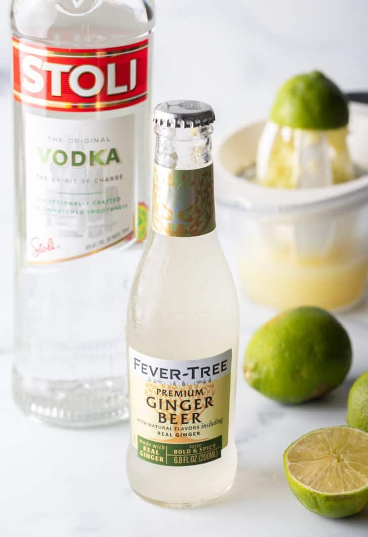 Ingredients you need: Glass bottle of vodka, bottle of ginger beer, whole fresh lime and in the background is half of a lime on a hand juicer.