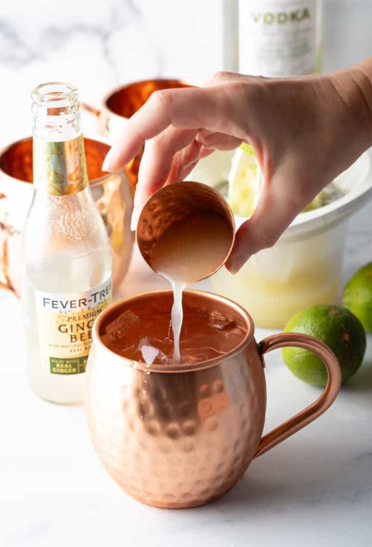 Hand pouring liquid from a small shot glass into a copper metal mug.