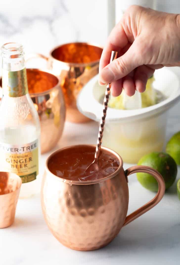 Hand using a metal stir stick to stir ginger beer and vodka in a copper metal mug.