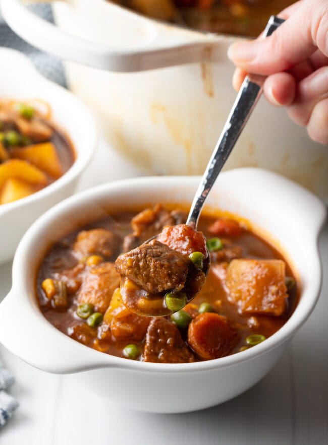 Mulligan beef stew with chopped potatoes, carrots, peas, and chunks of meat in a white bowl. Hand with metal spoon showing a spoonful to camera.