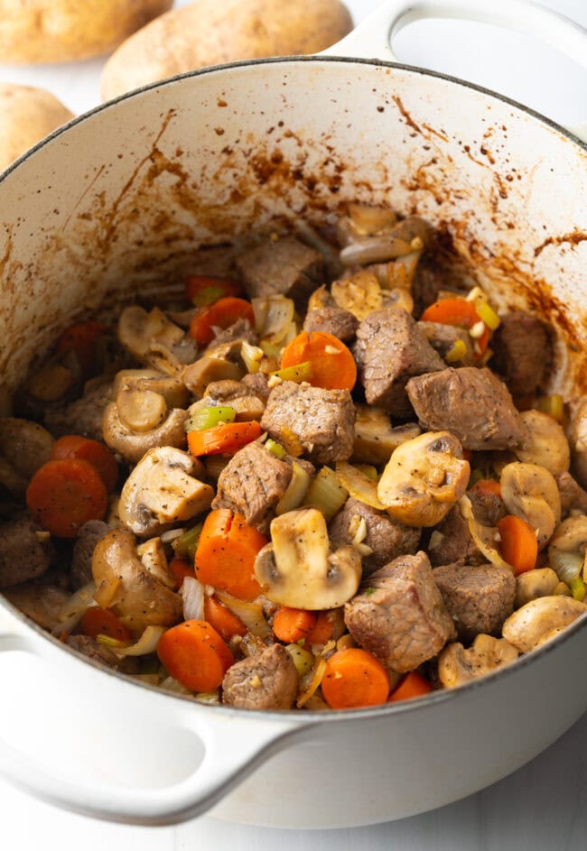 Chopped carrots, celery, onion, mushrooms, and potatoes with chunks of beef in a pot.