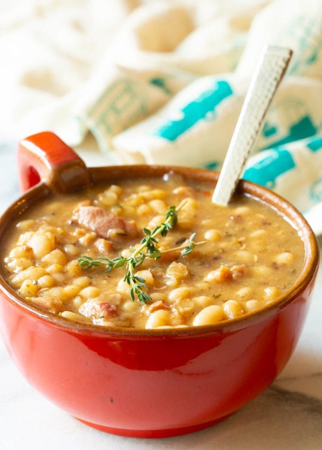 Single serving of navy bean soup with beans in a red bowl with a handle and a metal spoon sticking out 
