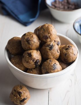 White bowl brimming with energy balls that are speckled with chocolate chips and raisins. One ball is on the table next to the bowl.