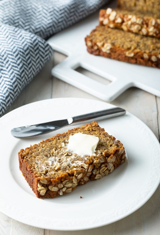 Slice of banana bread on a white plate with butter on it. 