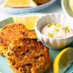 Two salmon patties on a green plate, with a white ramekin of tartar sauce and two lemon wedges.