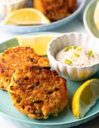 Two salmon patties on a green plate, with a white ramekin of tartar sauce and two lemon wedges.