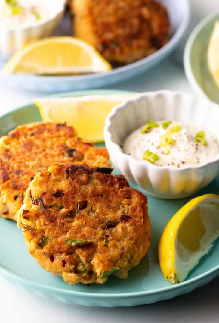 Two salmon patties on a green plate, with a white ramekin of tartar sauce and two lemon wedges.